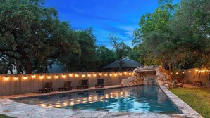 Un jardin avec une piscine, une bordure en pierre, des chaises longues en bois et des guirlandes lumineuses.