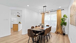Un espace repas moderne avec une table et des chaises en bois, une cuisine en arrière-plan et une grande fenêtre avec des rideaux.