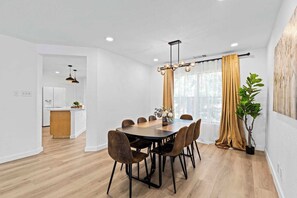 Ein moderner Essbereich mit Holztisch und Stühlen, eine Küche im Hintergrund und ein großes Fenster mit Vorhängen.