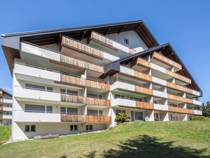 Um edifício de vários andares com varandas, um telhado inclinado e uma colina gramada ao fundo.