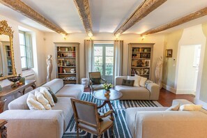 Ein geräumiges Wohnzimmer mit hohen Decken, sichtbaren Balken, einem runden Couchtisch und einem großen Fenster mit Vorhängen.