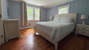 A bedroom with a bed, nightstands, a dresser, and two windows with curtains. - Charming 2 bedroom cottage by the pond in Eastham, Cape Cod (Eastham)