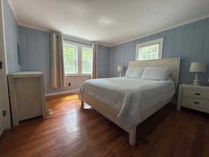 A bedroom with a bed, nightstands, a dresser, and two windows with curtains.