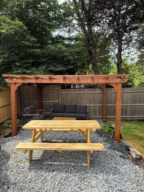 A wooden pergola with a bench and a table, a black outdoor sofa, and a wooden fence in the background.