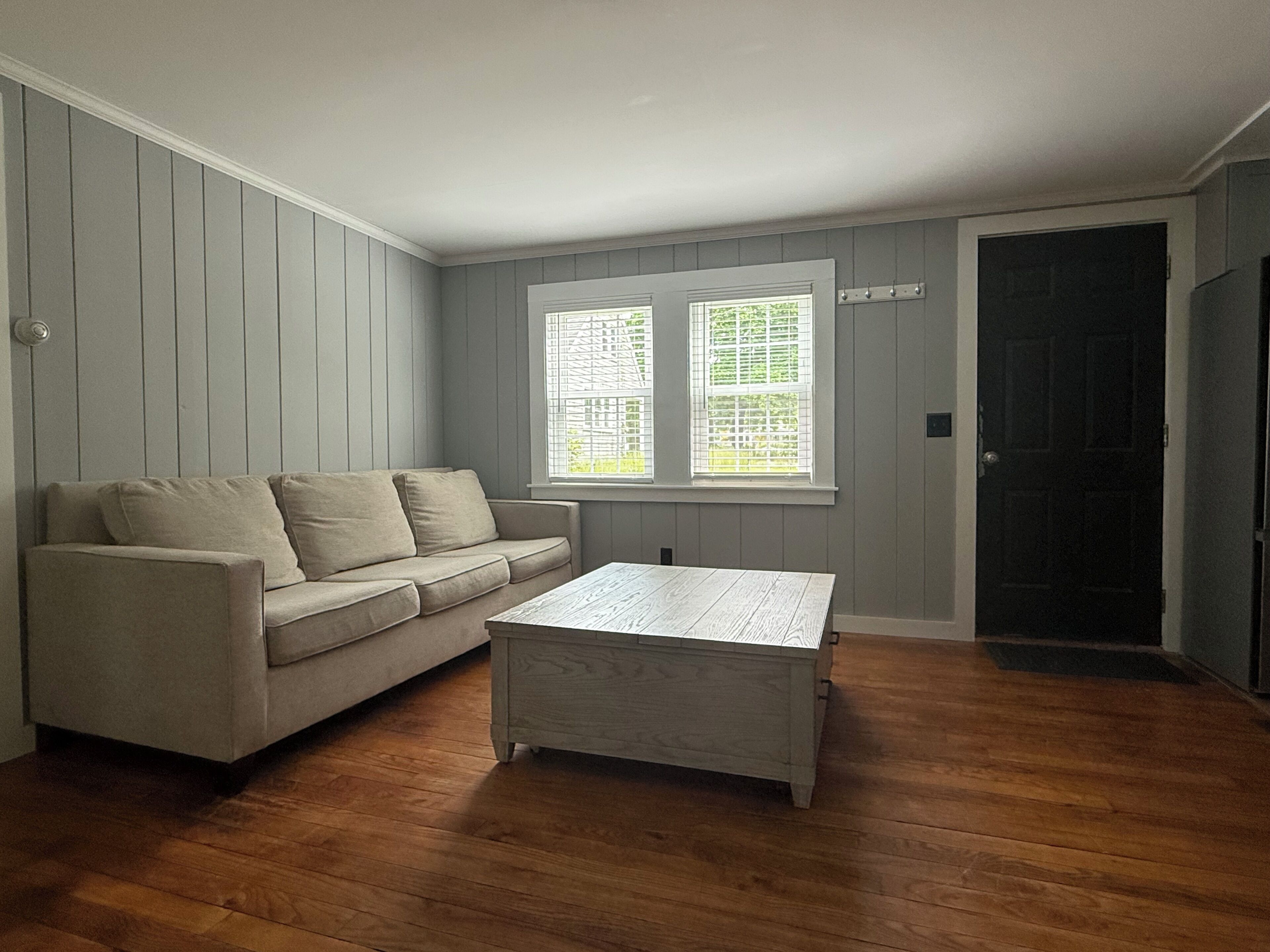 A room with a beige sofa, a wooden coffee table, a window with blinds, and a door.