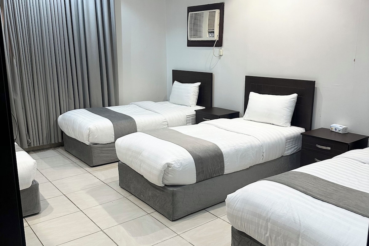 A hotel room with three beds, a large window with curtains, a wall-mounted air conditioner, and a tiled floor.