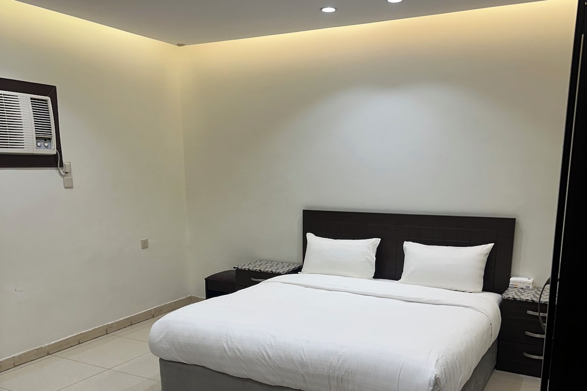 A modern bedroom with a large bed, bedside tables, a wall-mounted air conditioner, and a patterned rug.