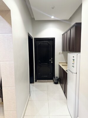 A narrow kitchen with a black door, a white refrigerator, and dark wooden cabinets. - Nour Al Raqia Suites (Madinah)