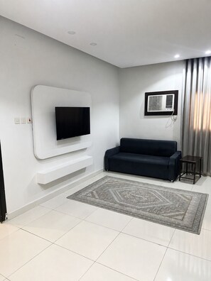 A modern living room with a flat-screen TV mounted on a white wall, a dark-colored sofa, a patterned rug, and a small table. - Nour Al Raqia Suites (Madinah)