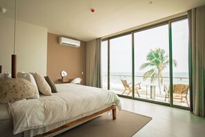 Une chambre à coucher avec un grand lit, une table de chevet, une chaise et une vue sur l’océan grâce à de grandes fenêtres.