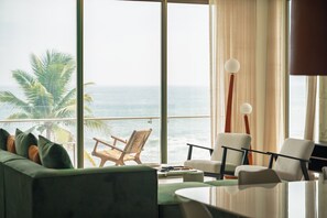 Ein Zimmer mit großem Fenster zum Meer, einem grünen Sofa, einem weißen Sessel und einem Tisch mit Lampe.
