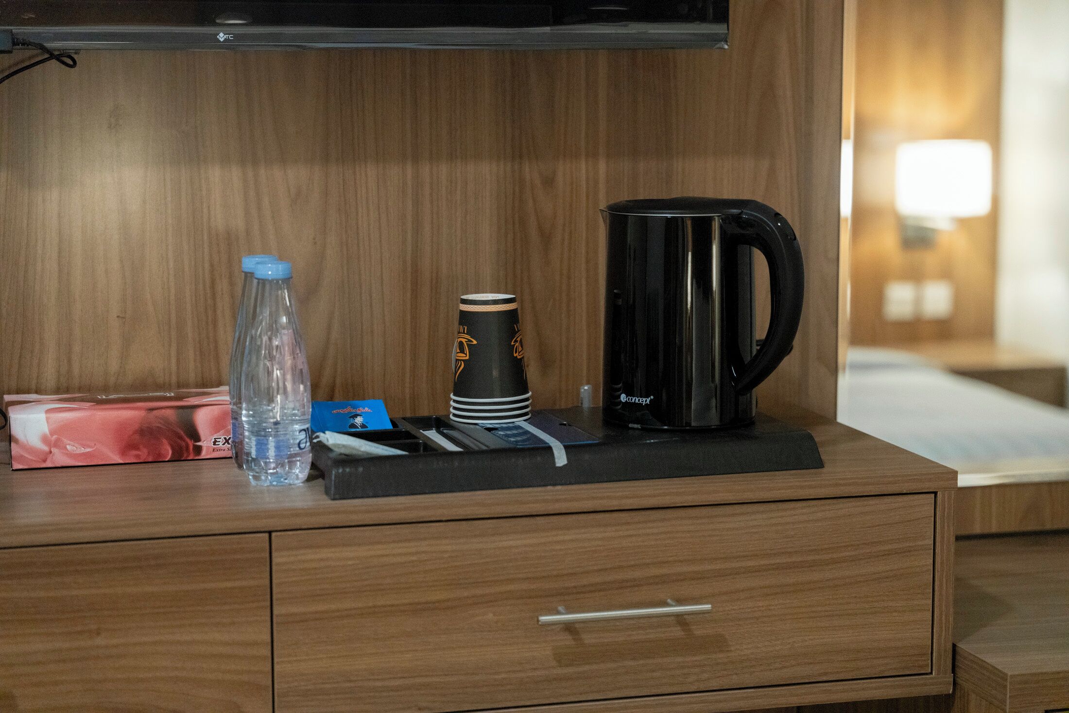 A wooden bedside table with a black coffee maker, a clear water bottle, a black tumbler, and a tissue box.