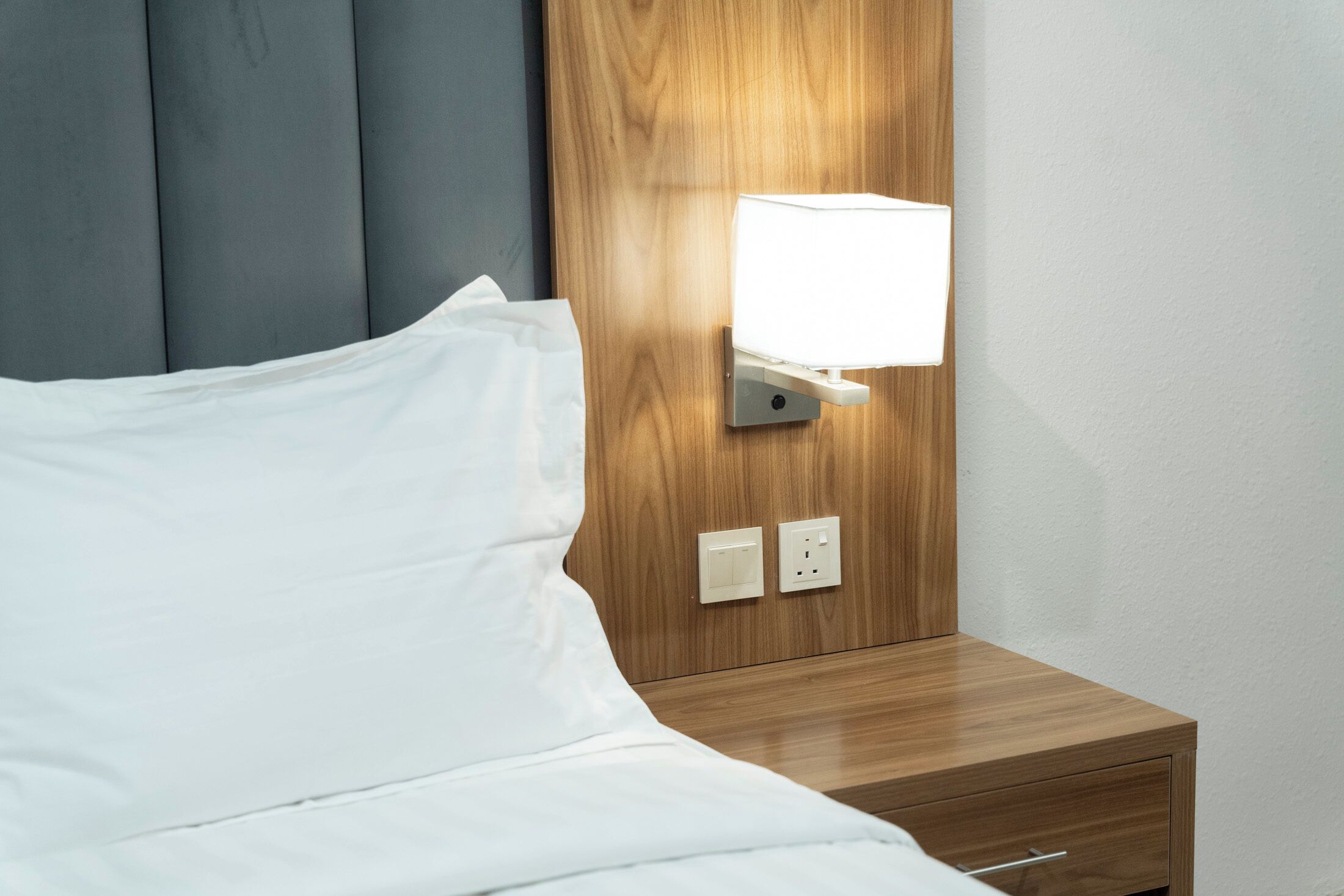 Una cama con ropa blanca, cabecera de madera, mesita de noche con lámpara y dos enchufes.