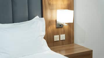 Una cama con ropa blanca, cabecera de madera, mesita de noche con lámpara y dos enchufes.