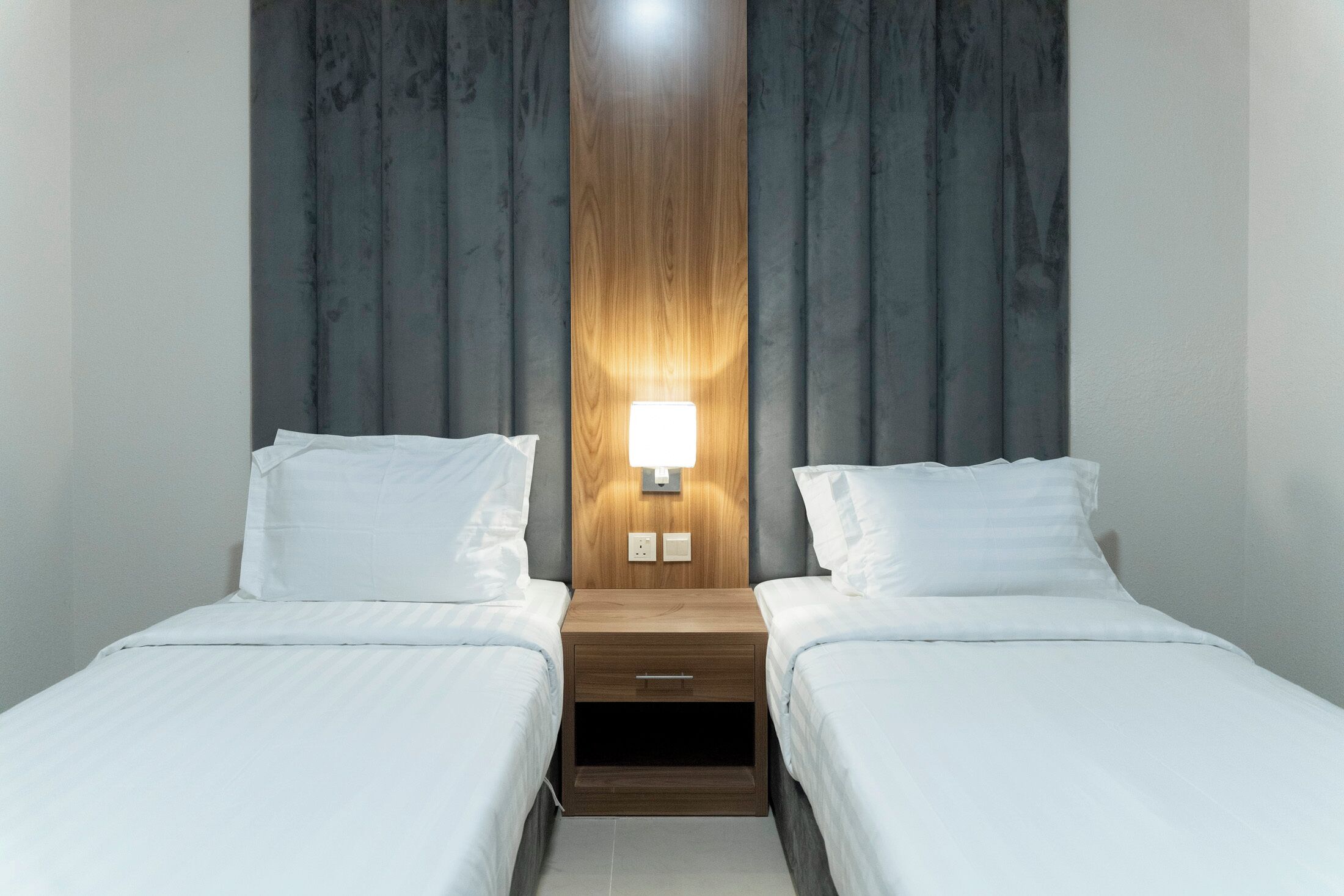 Habitación de hotel con dos camas, cabecero de madera, mesita de noche con lámpara y pared lisa.