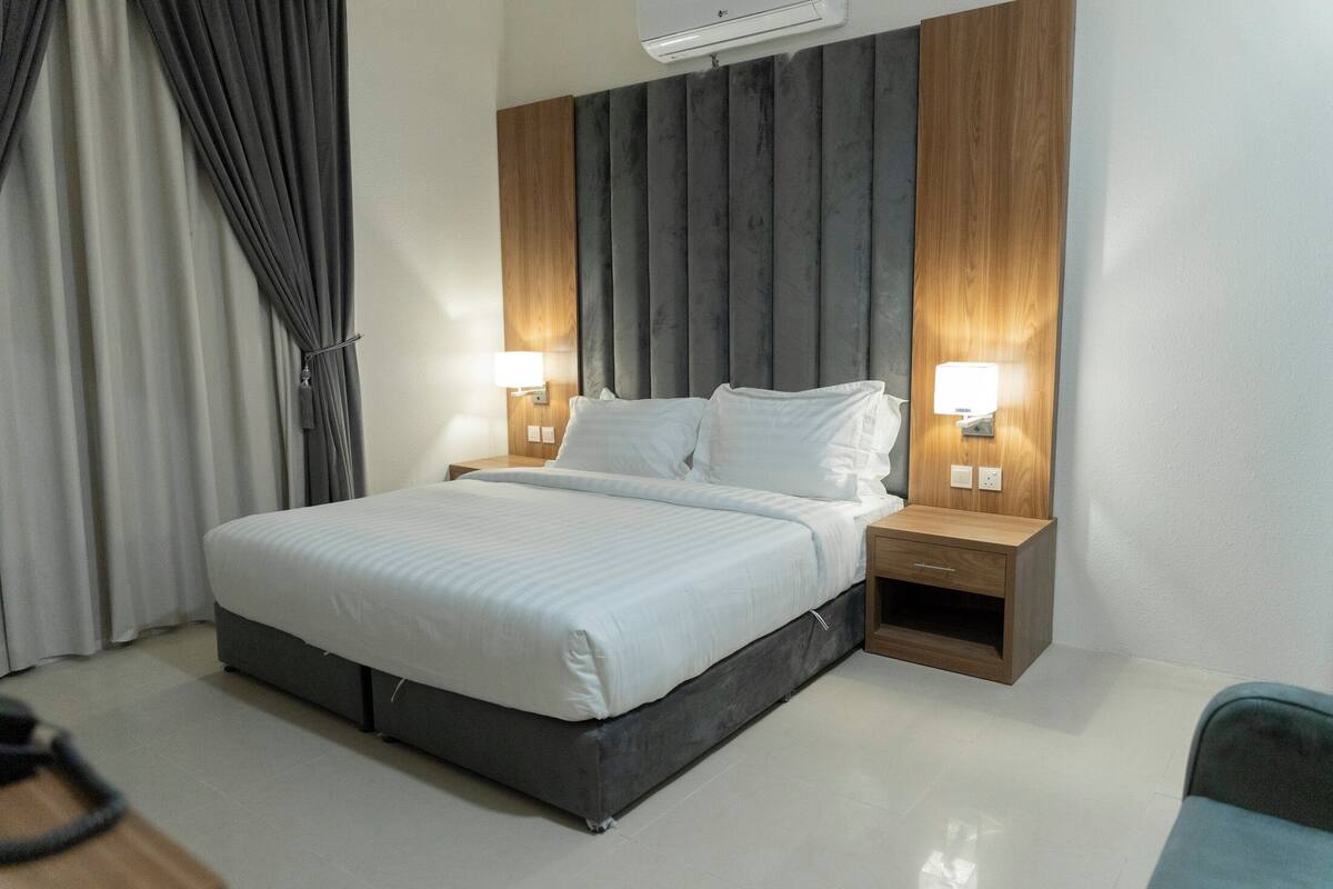 Una habitación de hotel moderna con una cama grande, dos mesitas de noche con lámparas, un cabecero de madera y una cortina gris.