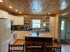 A kitchen with white cabinets, a wooden ceiling, a central island with stools, and a window with a view. - Camp Oliver Rose - Inlet's hidden gem (Inlet)
