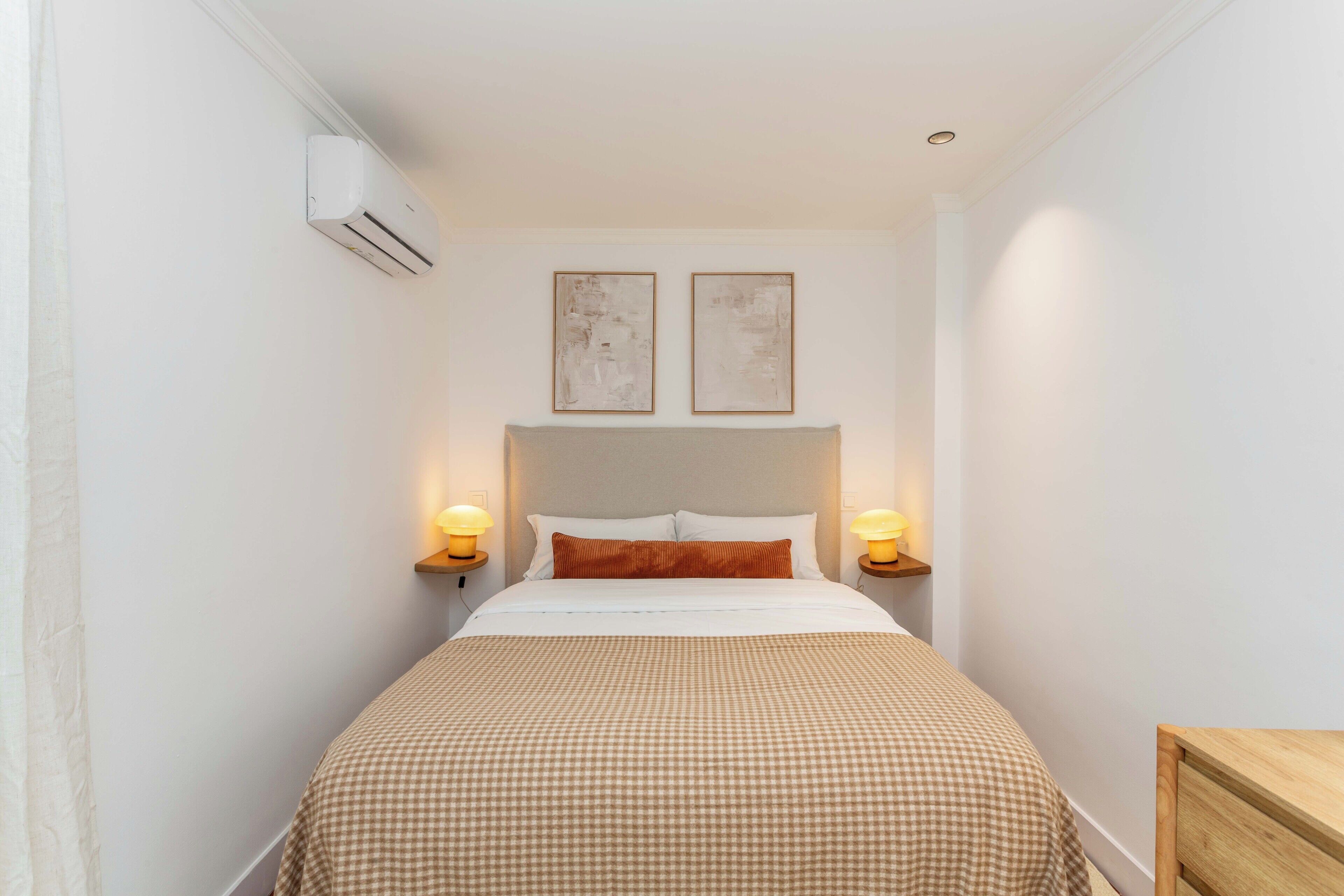 Ett litet hotellrum med en enkel säng, två väggmonterade luftkonditioneringsapparater och två sänglampor.