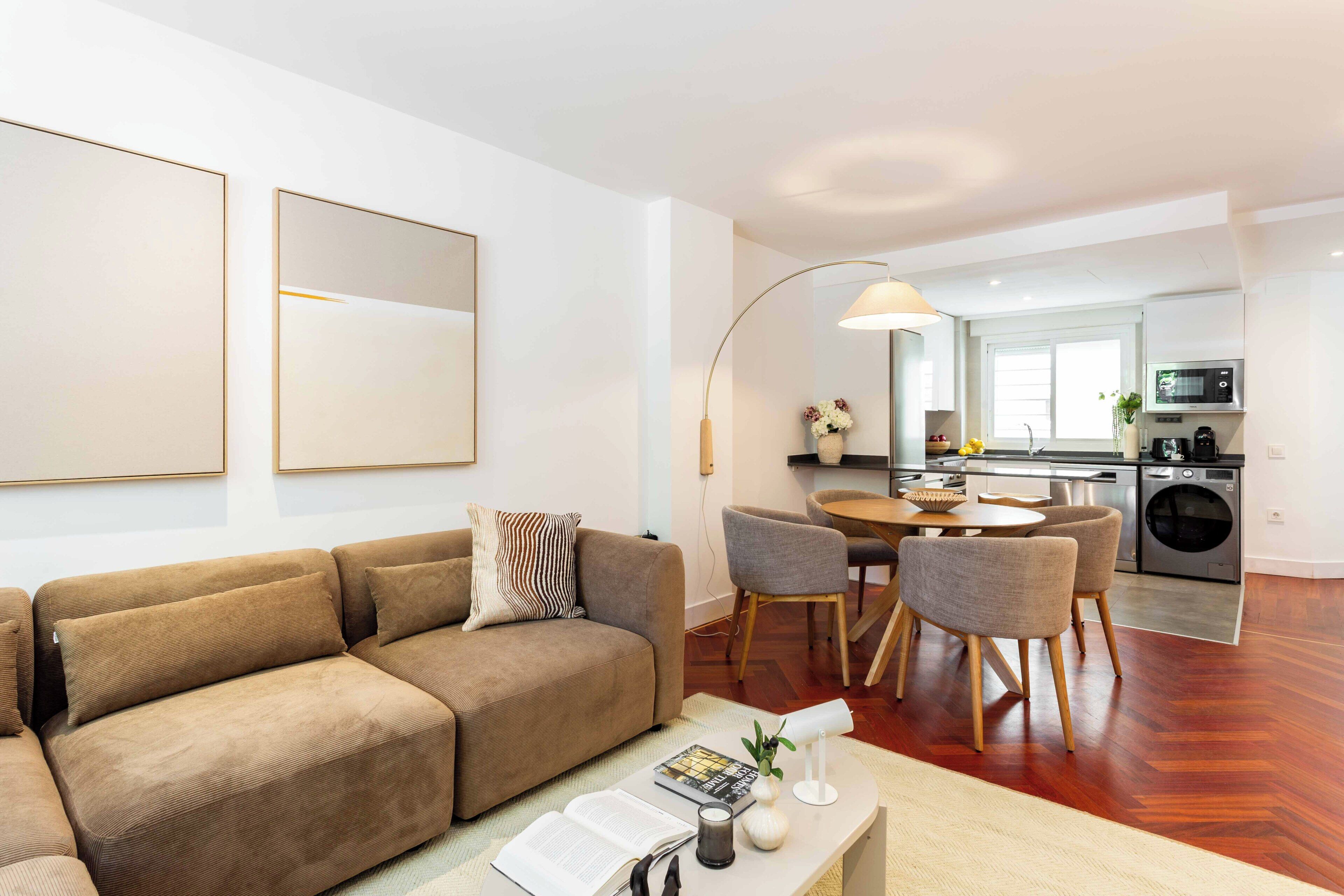 A modern living room with a sofa, a dining table with chairs, a kitchen area with appliances, and a large mirror.
