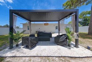 A covered outdoor seating area with a grill, two chairs, and a table, surrounded by gravel and plants. - Coastal haven in prime location (Clearwater)