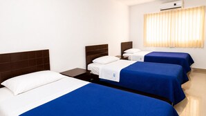 Habitación de hotel con tres camas, una mesita de noche, aire acondicionado en la pared y una ventana con cortinas.