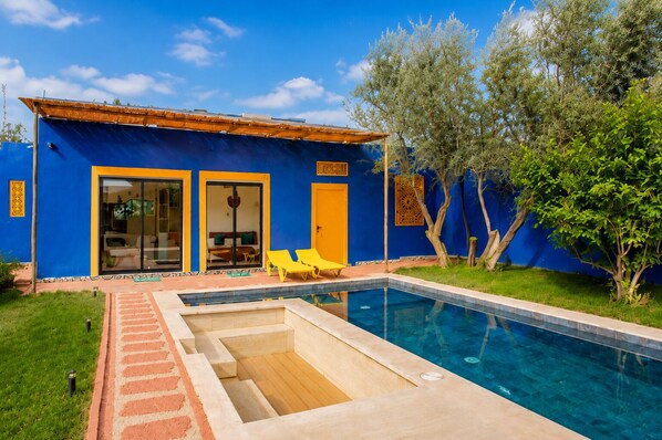 Un bâtiment bleu avec une piscine, deux portes et deux fauteuils de jardin jaunes.