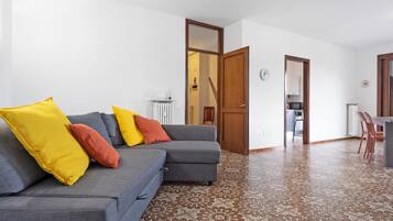 Un soggiorno con un divano grigio, cuscini gialli e rossi, un pavimento decorato e una porta in legno che dà su una cucina.