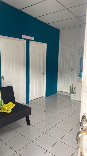 Une pièce avec deux portes blanches, un mur bleu, un sol carrelé blanc, un canapé noir avec un coussin jaune et une horloge au mur.