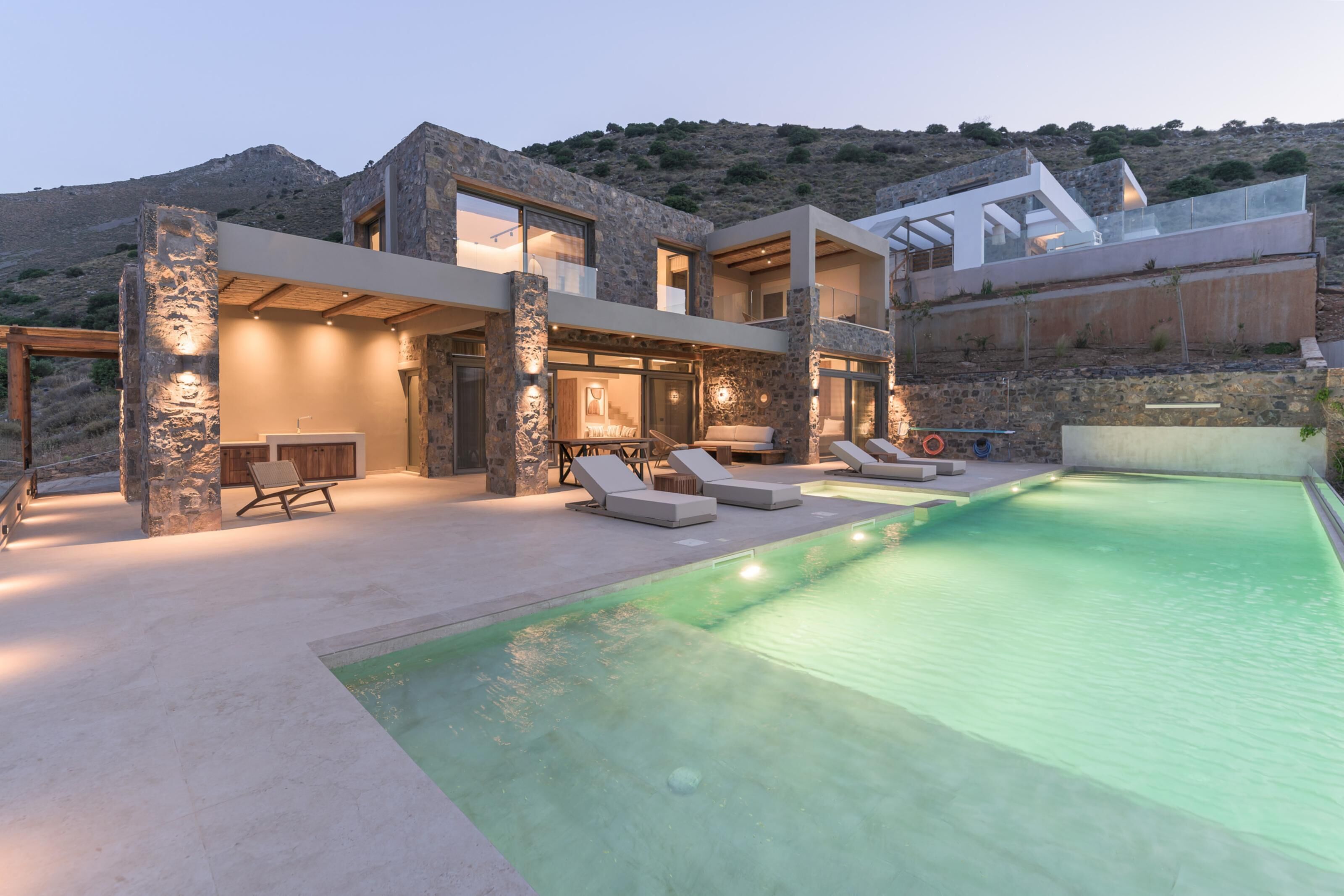 A modern house with a swimming pool, stone pillars, and a view of the hills.