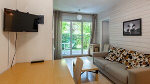 A modern living room with a flat-screen TV, a wooden dining table, a sofa with a cow print cushion, and a sliding glass door leading to a garden. - Bright apartment with nature view 4 people (Ax-les-Thermes)