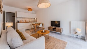 A modern living room with a sofa, coffee table, TV, and kitchen area. - “Cocon Des Sables-Bassin d’Arcachon” Apartment with Shared Pool, Private Terrace & Wi-Fi (La Teste-de-Buch)