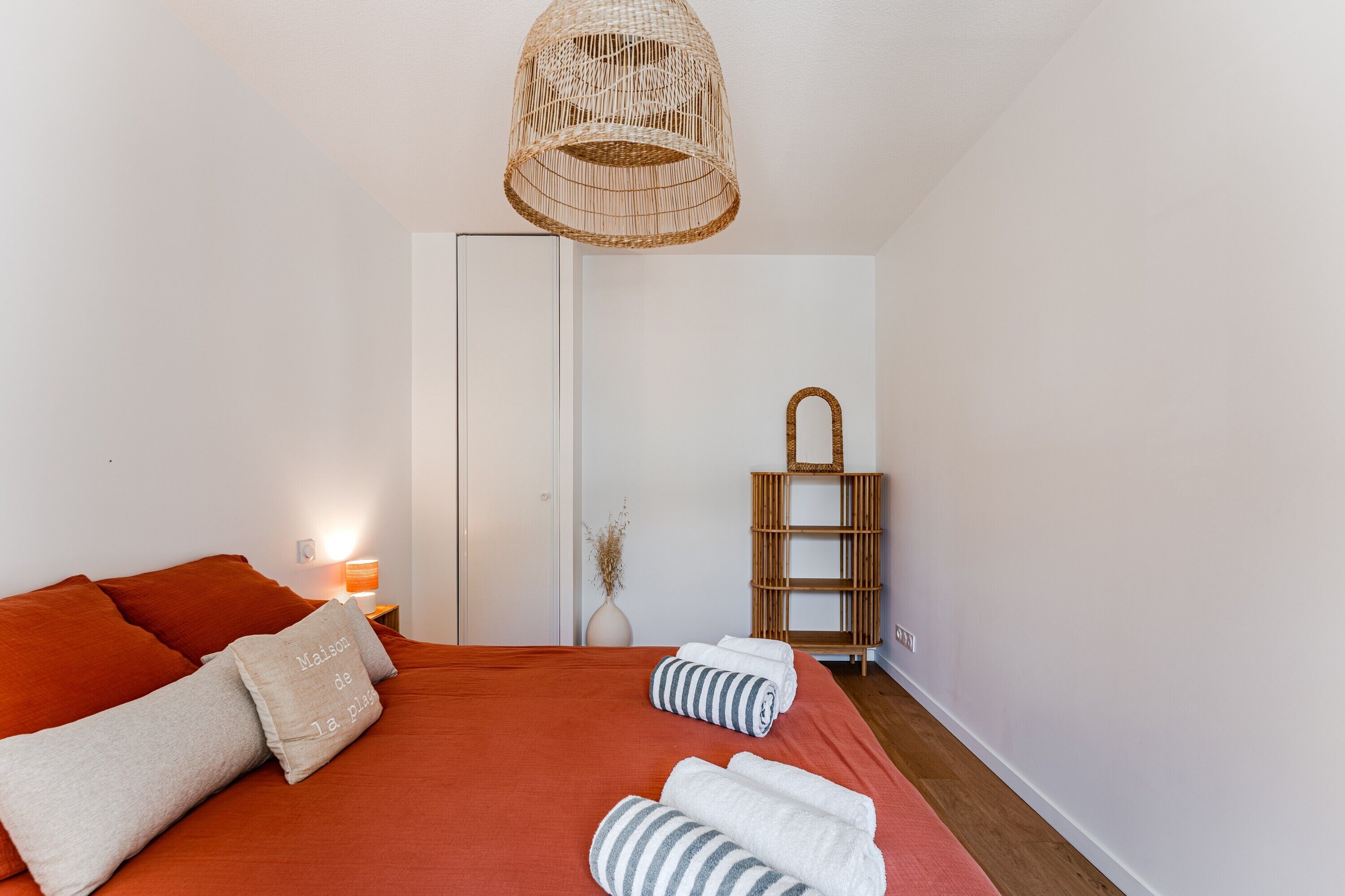Un dormitorio con cama, almohadas, una lámpara de noche, un colgante de luz de mimbre, un estante de madera y una puerta.