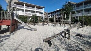 A beachside playground with a slide and climbing structures, surrounded by buildings with balconies. - “Cocon Des Sables-Bassin d’Arcachon” Apartment with Shared Pool, Private Terrace & Wi-Fi (La Teste-de-Buch)