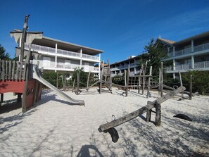 Un parque infantil junto a la playa con un tobogán y estructuras para escalar, rodeado de edificios con balcones.