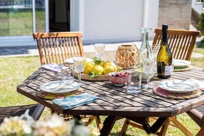 Una mesa de madera al aire libre con platos, vasos, una botella y un cuenco de fruta.