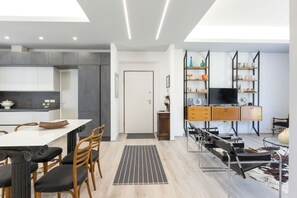 Una cocina moderna con armarios grises, un comedor con mesa y sillas blancas, y una sala de estar con televisor y estanterías.