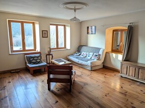 Ein Zimmer mit Holzboden, einer Couch, einem Sessel, einem Tisch und zwei Fenstern.