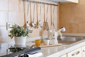 Una cocina con encimera blanca, cafetera, planta y utensilios colgando en la pared.