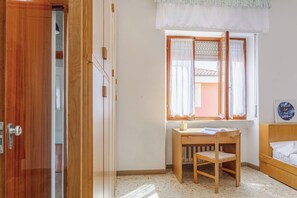 Um quarto com porta de madeira, janela com cortinas, escrivaninha de madeira e uma cama.