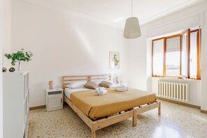 Una camera da letto con un letto di legno, pareti bianche, un termosifone, una finestra con tende e un comodino con una lampada, oltre a un quadro appeso al muro.