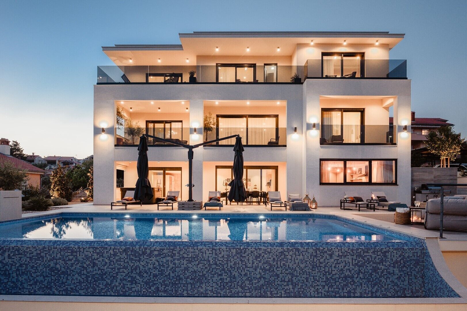 Una casa moderna de varios pisos con piscina, mobiliario exterior y una entrada bien iluminada.