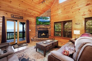 L’intérieur chaleureux d’un chalet avec une cheminée, des murs en bois, un canapé et une vue sur le paysage à travers la porte.