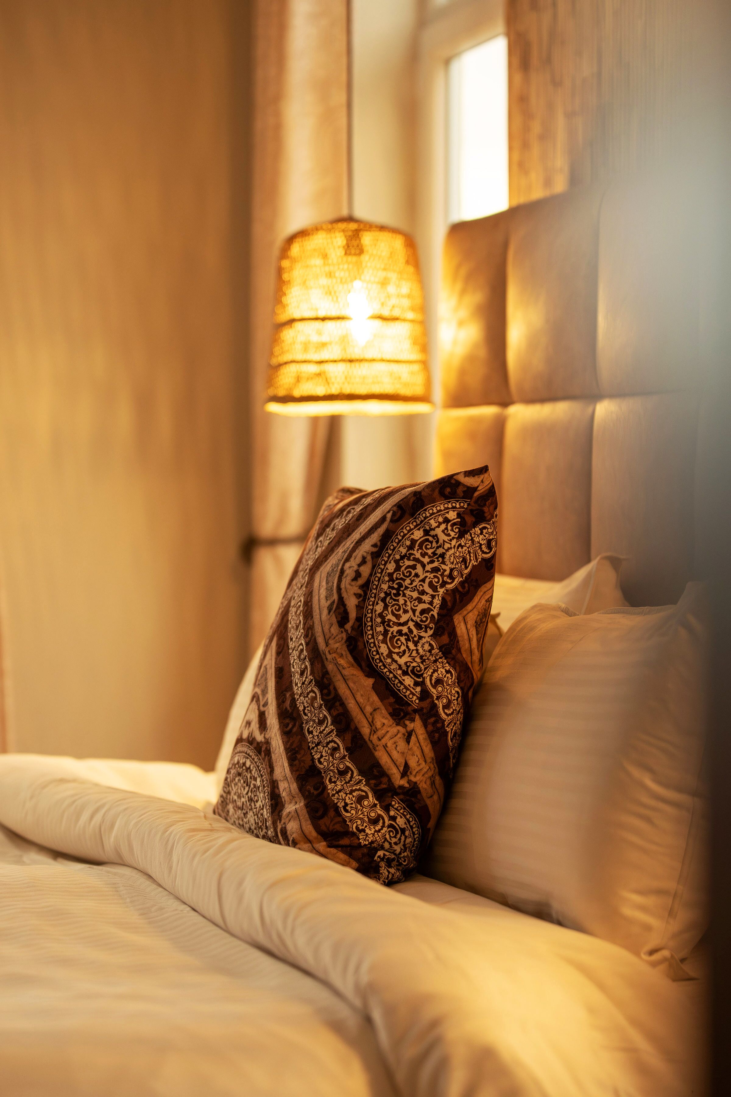 Una cama con una almohada estampada, una lámpara colgante y una ventana con cortinas.