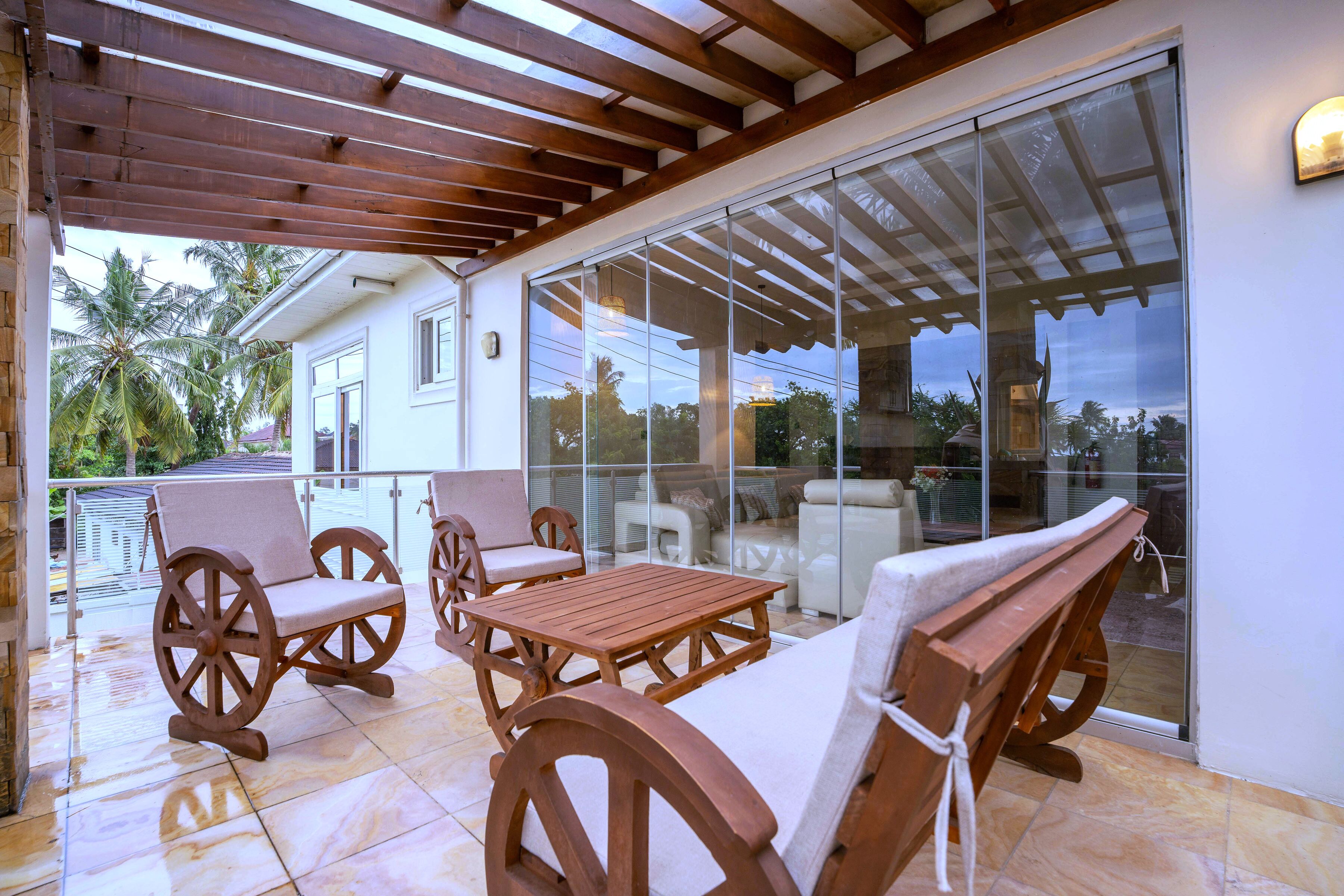 Un patio cubierto con mobiliario de madera, una puerta de cristal que da a una piscina y una casa blanca al fondo.