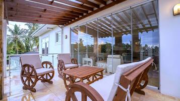 Un patio cubierto con mobiliario de madera, una puerta de cristal que da a una piscina y una casa blanca al fondo.
