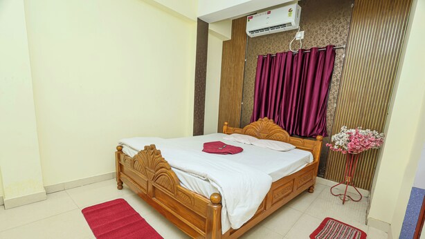 A neatly arranged bedroom with a wooden bed, a red rug, a small table with a vase, and a curtain. - Silver Residency (Bhubaneshwar)