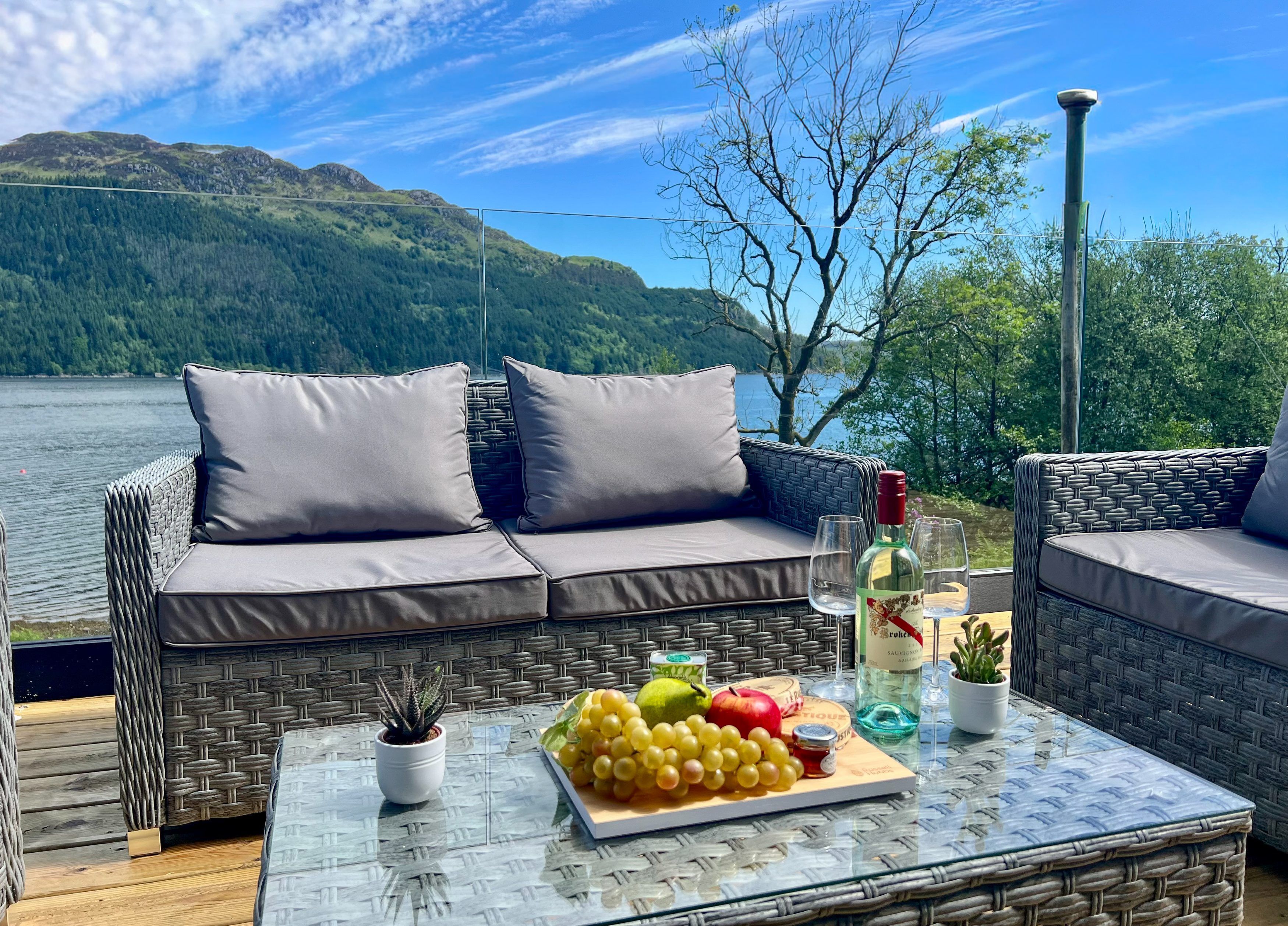 Un canapé en osier avec des coussins, une table en verre avec des fruits et une bouteille, et une vue sur un lac et des montagnes.