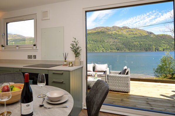 Una cocina moderna con vistas a un lago y montañas, un comedor con mesa para dos, y un balcón con mobiliario de exterior.