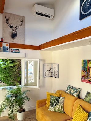 Un salon avec un canapé, une plante en pot, des œuvres d’art mural et une fenêtre donnant sur un beau paysage.
