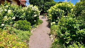 Un sendero en un jardín que conduce a un edificio de techo rojo, rodeado de vegetación exuberante y plantas con flores.